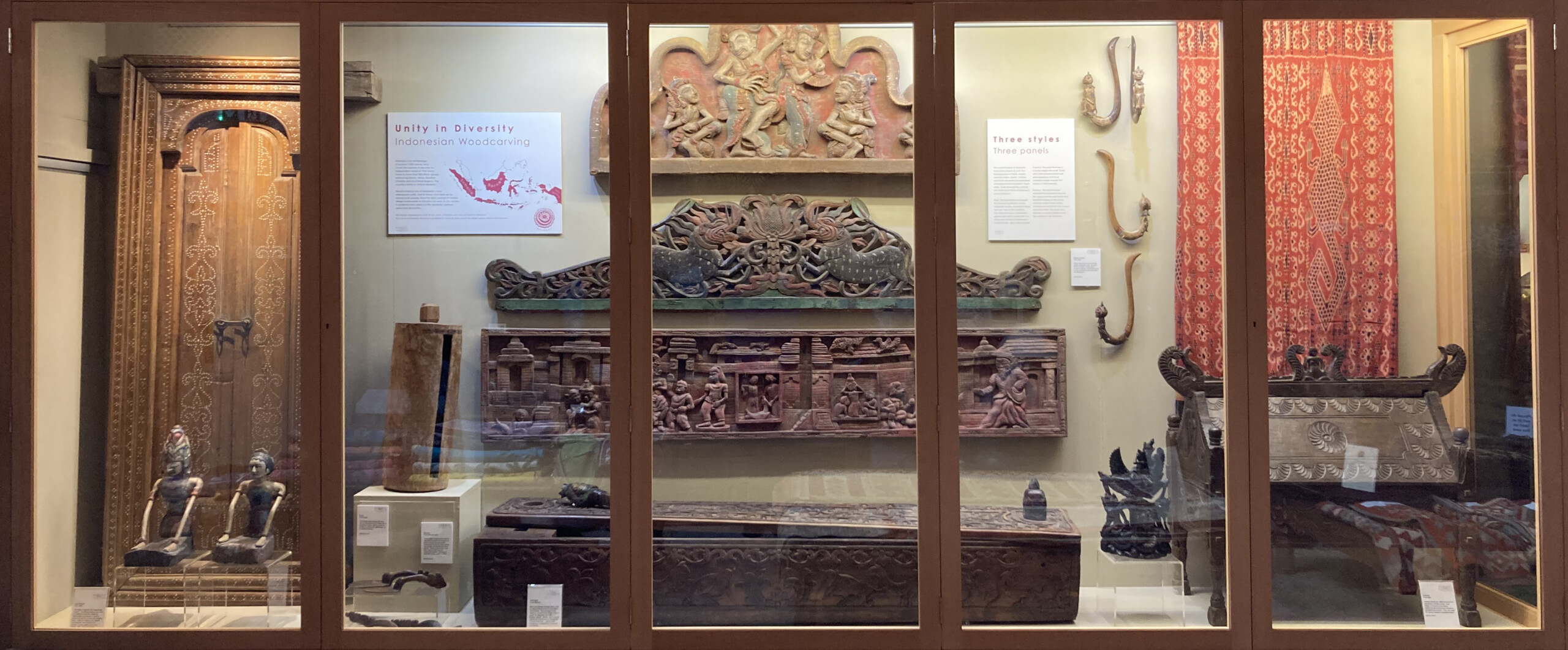 This shows the curated display case with lots of wood carved objects and textiles with some interpretation boards and labels.