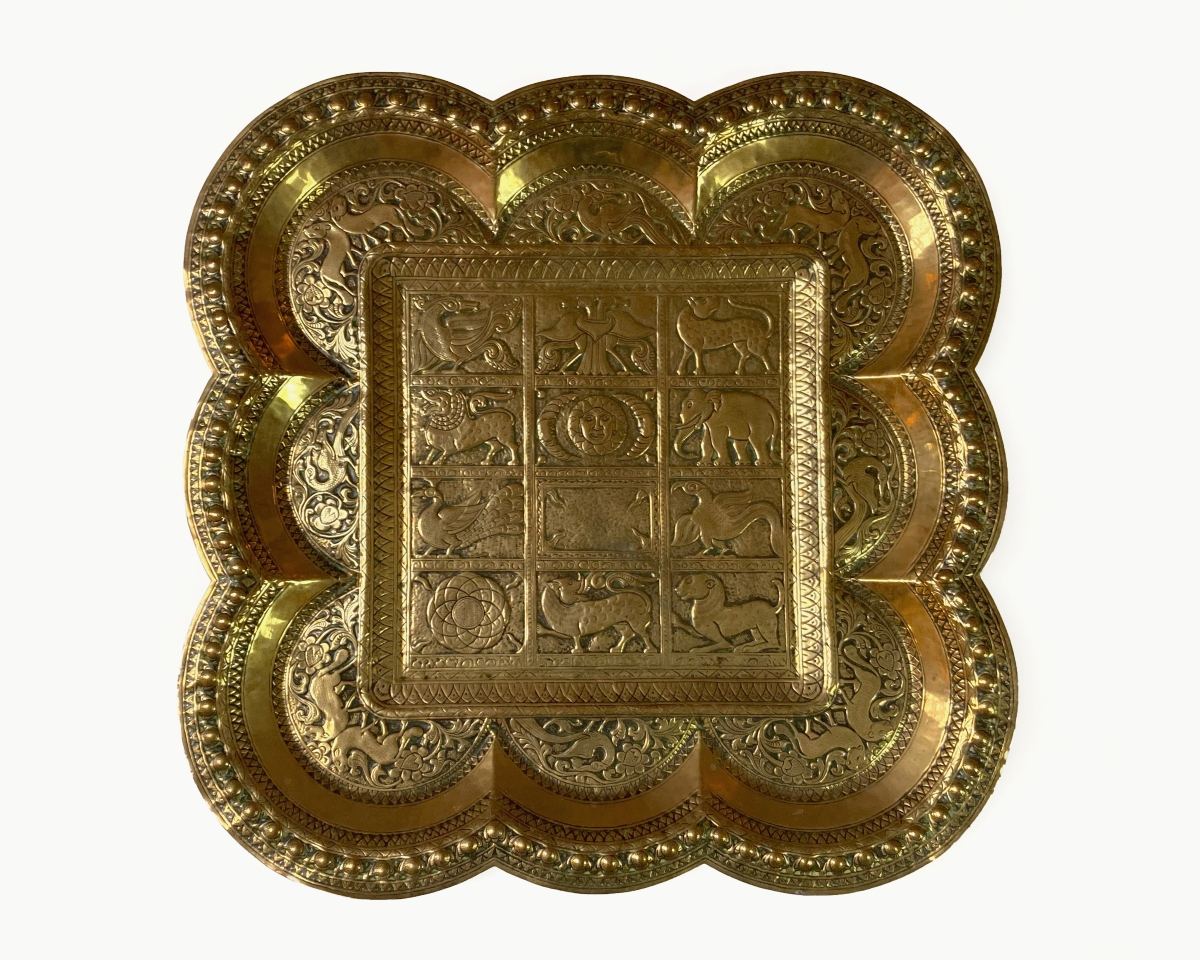 An intricately designed metal tray on a white background. It is brass which is a dull golden colour and a bit shiny. The central part of the tray is a square with 12 rectangular sections in a grid with an ornate border. The outer part of the tray is 3 semi-circle shapes coming out of each side, making the silhouette have a cartoon cloud like shape. The Rectangular sections show symbols such as various animals, the sun and moon, and some more geometric and minimal ones.