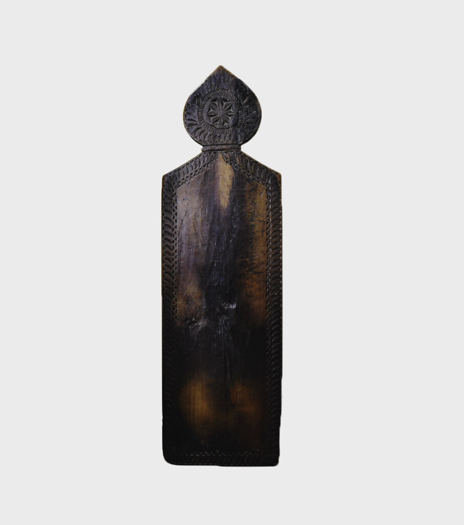 A vertical wooden prayer board. It is made of dark wood and you can see lighter patches where people have knelt and it is worn down. It is a rectangle with a rounded pointed part on the rop. There is some ornate carving in the top bit.