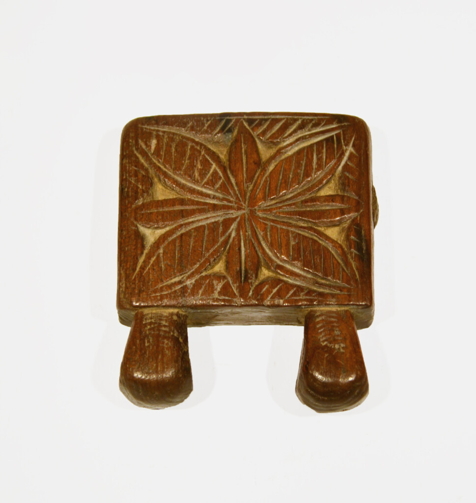 This is a small square wooden object with a geometric carving in the centre. It has two small parts jutting out at the bottom presumably to attach it to something.