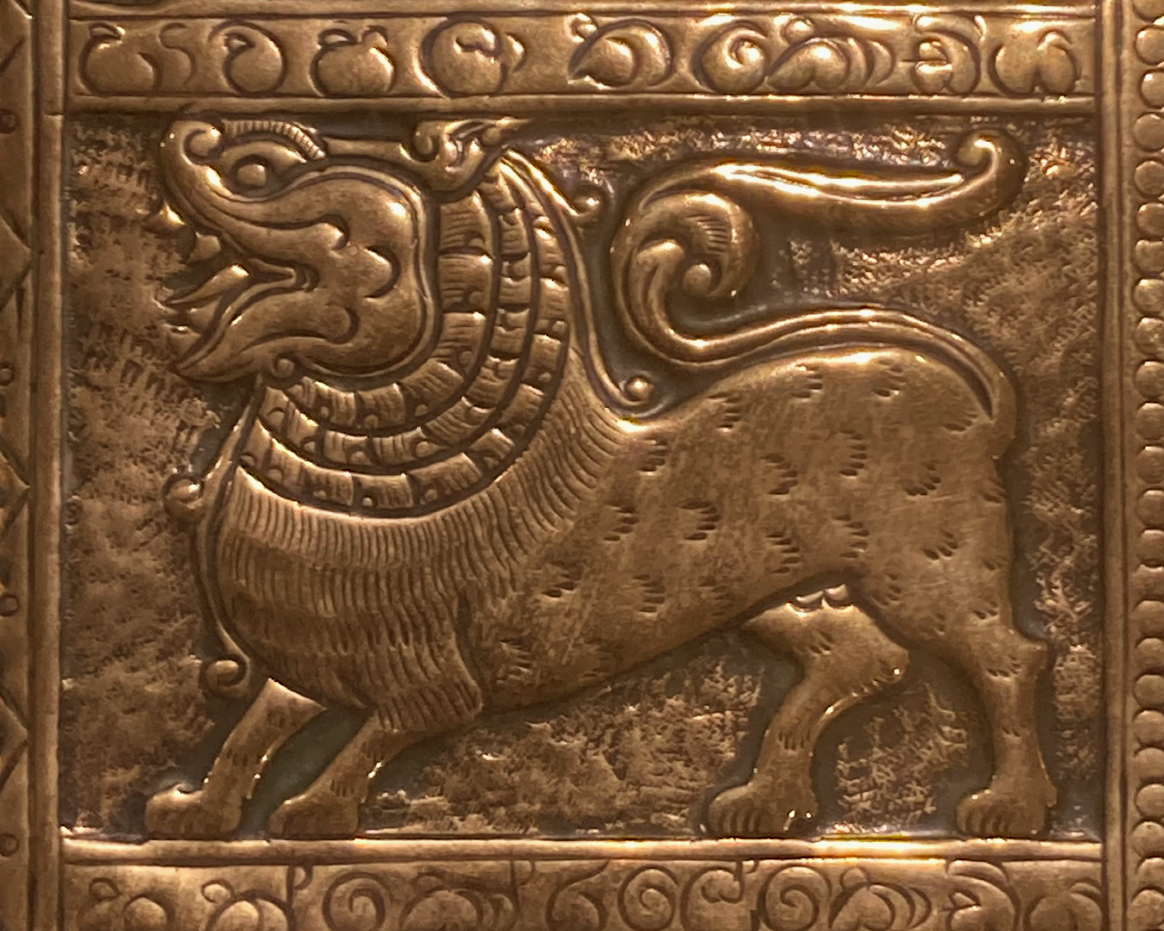 This shows part of the tray, it is a side profile of a stylised lion with its mouth open, a long tail and a big mane. There is an ornate border around this.