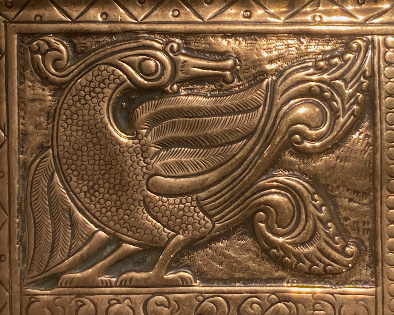 This is a close up of the tray. It shows a the side profile of a stylised swan, it's head is facing backwards and it has a wing raised and elaborate tail feathers. It is ornately detailed. There is a border to this image with some pattern.